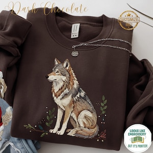May include: A dark chocolate-coloured sweatshirt with an embroidered wolf design. The wolf is in shades of grey, brown, and beige, with green foliage accents. The text "Dark Chocolate" is at the top. The sweatshirt has a round neckline.
