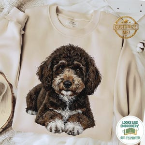 Puede incluir: Una sudadera beige con una ilustración detallada de un perro marrón y blanco. El perro tiene pelaje esponjoso y está acostado. La sudadera tiene cuello redondo y mangas largas. La imagen también incluye una etiqueta "Customize Me!"