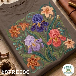 Puede incluir: Una camiseta marrón oscuro con un estampado floral detallado de lirios naranjas, iris morados e iris rosas con tallos y hojas verdes. Una insignia blanca circular en la esquina contiene el texto: LOOKS LIKE EMBROIDERY BUT IT'S PRINTED!