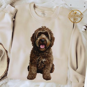 Puede incluir: Sudadera beige con una ilustración detallada de un perro Goldendoodle marrón. El perro está centrado en la parte delantera de la sudadera. Una pegatina dorada con las palabras "Customize Me!" es visible.