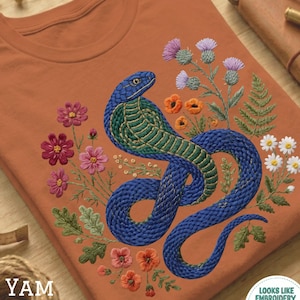 Op de afbeelding: Terracotta-kleurig T-shirt met een geborduurd ontwerp van een blauwe en groene slang te midden van kleurrijke bloemen en bladeren. Het ontwerp bevat roze, oranje en witte bloemen, met de tekst "YAM" linksonder.