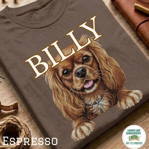 以下が含まれることがあります： 茶色のTシャツで、キャバリア・キング・チャールズ・スパニエルのグラフィックイラストが描かれています。犬の上には大きな白い文字でBILLYとプリントされています。犬は長くウェーブのかかった茶色の毛、黒い鼻、ピンクの舌が特徴です。