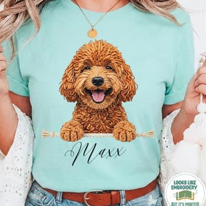 Op de afbeelding: Lichtblauw t-shirt met een afbeelding van een lachende bruine doodle hond. De hond houdt een touw vast met de naam "Maxx" eronder. Het shirt heeft ook de woorden "Chalky Mint" aan de onderkant.