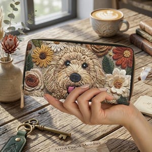 May include: A beige zip-around wallet featuring an embroidered image of a dog's face surrounded by colorful flowers. The wallet has a gold zipper pull and is held in a person's hand. The background includes a coffee cup and a vase with flowers.