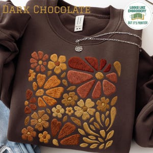 May include: A dark chocolate brown crewneck sweatshirt featuring a printed graphic of various flowers in shades of orange, rust, and gold. The text "DARK CHOCOLATE" appears in the top left, and a circular badge reads "LOOKS LIKE EMBROIDERY BUT IT'S PRINTED!" in the top right corner.