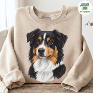 Könnte beinhalten: Ein beigefarbenes Sweatshirt mit Rundhalsausschnitt und einem mittigen, detaillierten Grafikdruck eines Australian Shepherd Hundes. Der Hund hat schwarzes, lohfarbenes und weißes Fell mit einer weißen Blesse im Gesicht. Ein rundes Abzeichen zeigt LOOKS LIKE EMBROIDERY BUT IT'S PRINTED.