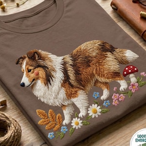 May include: A brown t-shirt featuring an embroidered Shetland Sheepdog with brown, white, and tan fur. The dog is surrounded by embroidered flowers, leaves, and a mushroom. The shirt is on a wooden surface.