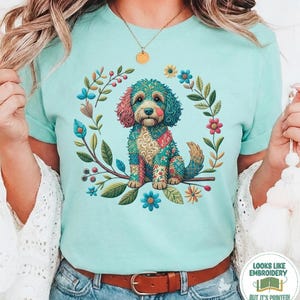 Puede incluir: Una camiseta de color verde menta pálido con un gráfico central de un perro con un patrón de mosaico colorido e intrincado. El perro está rodeado por una corona circular de varias flores y hojas en tonos azules, rosas, amarillos y verdes.