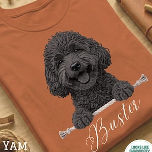 Può includere: T-shirt color terracotta con una grafica di un cane nero sorridente che tiene una corda bianca, con il nome "Buster" in corsivo bianco. La maglietta ha anche il testo "YAM" e un logo che dice "Looks Like Embroidery But It's Printed!"