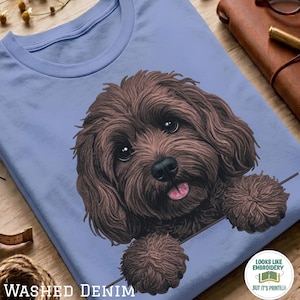 Può includere: Una t-shirt azzurro chiaro con un'illustrazione grafica di un cane marrone dal pelo riccio, con occhi scuri e lingua rosa. Il cane sembra appoggiare le zampe su una linea orizzontale. La maglietta presenta un girocollo e maniche corte.