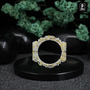 May include: A square-shaped, silver and gold-toned bracelet with a repeating cross design. The bracelet is encrusted with small, sparkling stones. The jewelry is displayed on a black pedestal against a blurred background.