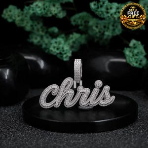 May include: A silver-toned pendant featuring the name "chris" in a cursive script font, entirely encrusted with small, clear, sparkling stones. The pendant includes a rectangular bail at the top, also covered in the same clear stones.