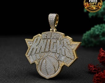 Peut inclure: Un pendentif doré représentant le logo des New York Knicks, entièrement incrusté de petites pierres claires. Le design comprend le nom de l'équipe en lettres stylisées au-dessus d'un ballon de basket, avec une bélière sertie de pierres.