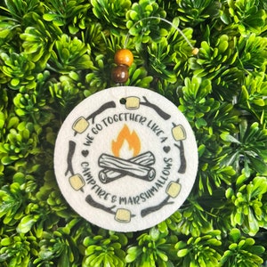 May include: White round felt ornament with a black and white graphic of a campfire and the text "We go together like a campfire & marshmallows".