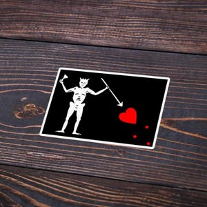 May include: A rectangular sticker featuring a black background with a white skeleton figure holding a spear and a red heart. The sticker has a white border and is placed on a dark wooden surface.
