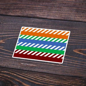 May include: A rectangular sticker with horizontal stripes in orange, blue, green, and maroon, each with white diagonal stripes. The sticker is on a dark wood surface.