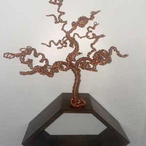 May include: A copper wire tree sculpture with a twisted, branch-like design, mounted on a hexagonal wooden shelf.