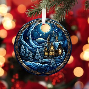 May include: A round Christmas ornament with a blue and gold design. The ornament features a snowy winter scene with a house, trees, and a full moon in the sky.