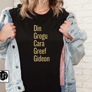 May include: Black t-shirt with gold text that reads "Din Grogu Cara Greef Gideon".