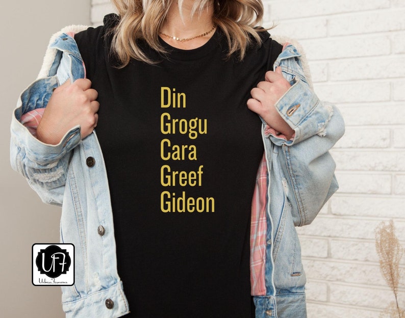 May include: Black t-shirt with gold text that reads "Din Grogu Cara Greef Gideon".