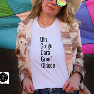 May include: White t-shirt with the text "Din Grogu Cara Greef Gideon" printed on it. The shirt is being worn by a person with a brown and white striped shirt over it.
