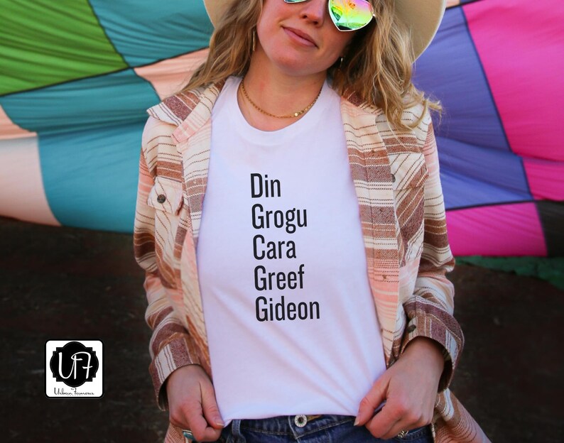 May include: White t-shirt with the text "Din Grogu Cara Greef Gideon" printed on it. The shirt is being worn by a person with a brown and white striped shirt over it.