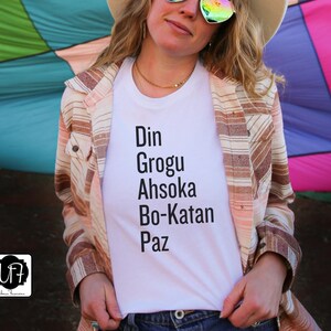 Puede incluir: Una camiseta blanca con el texto "Din Grogu Ahsoka Bo-Katan Paz" impreso en ella. La camiseta la lleva puesta una persona con una camisa de rayas marrones y blancas encima.
