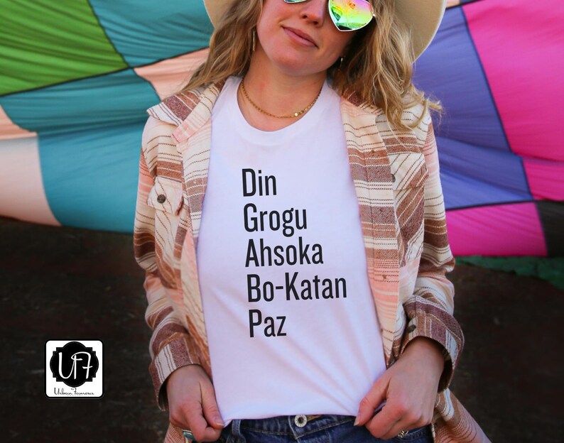 Puede incluir: Una camiseta blanca con el texto "Din Grogu Ahsoka Bo-Katan Paz" impreso en ella. La camiseta la lleva puesta una persona con una camisa de rayas marrones y blancas encima.