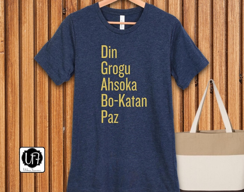 Puede incluir: Una camiseta azul marino con los nombres Din, Grogu, Ahsoka, Bo-Katan y Paz impresos en letras doradas.