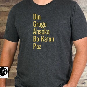 Puede incluir: Una camiseta gris oscuro con los nombres Din, Grogu, Ahsoka, Bo-Katan y Paz impresos en letras doradas.