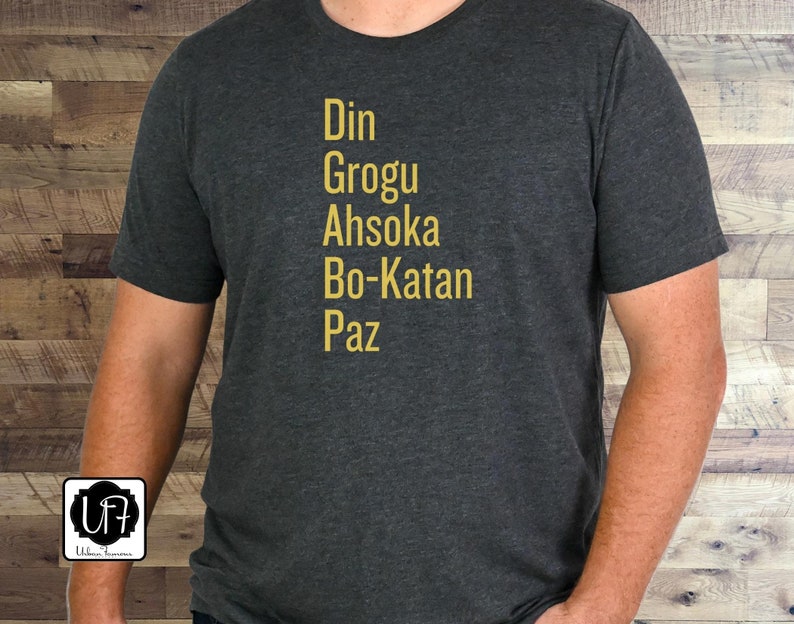 Puede incluir: Una camiseta gris oscuro con los nombres Din, Grogu, Ahsoka, Bo-Katan y Paz impresos en letras doradas.