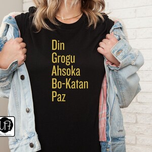 Puede incluir: Camiseta negra con texto dorado que dice "Din Grogu Ahsoka Bo-Katan Paz"