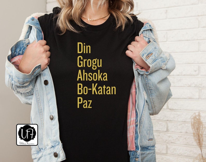 Puede incluir: Camiseta negra con texto dorado que dice "Din Grogu Ahsoka Bo-Katan Paz"
