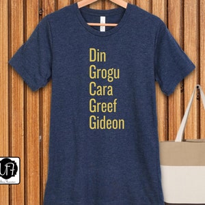 May include: A navy blue t-shirt with the names Din, Grogu, Cara, Greef, and Gideon printed in yellow on the front.