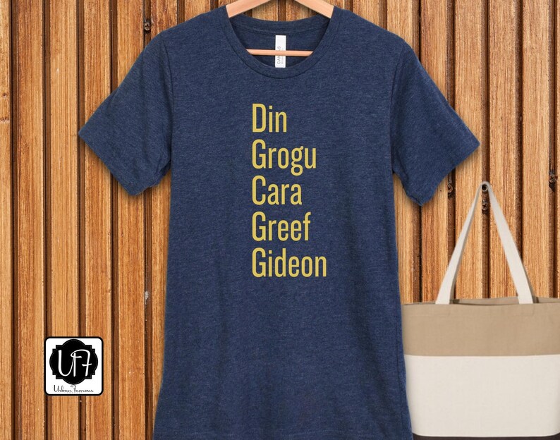 May include: A navy blue t-shirt with the names Din, Grogu, Cara, Greef, and Gideon printed in yellow on the front.