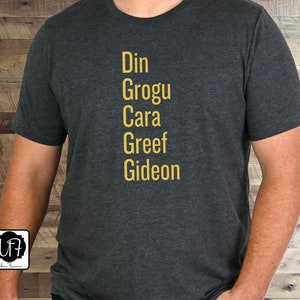 May include: A dark gray t-shirt with the names Din, Grogu, Cara, Greef, and Gideon printed in yellow on the front.