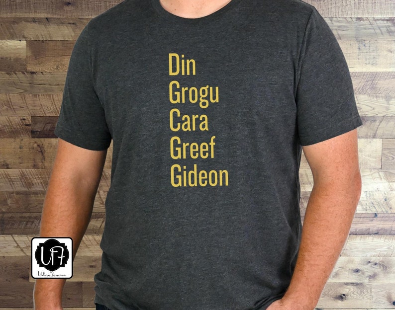 May include: A dark gray t-shirt with the names Din, Grogu, Cara, Greef, and Gideon printed in yellow on the front.