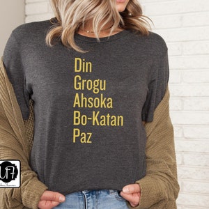 Puede incluir: Una camiseta gris oscuro con las palabras "Din Grogu Ahsoka Bo-Katan Paz" impresas en amarillo.
