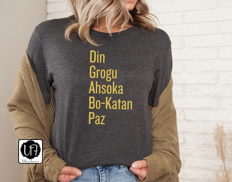 Puede incluir: Una camiseta gris oscuro con las palabras "Din Grogu Ahsoka Bo-Katan Paz" impresas en amarillo.