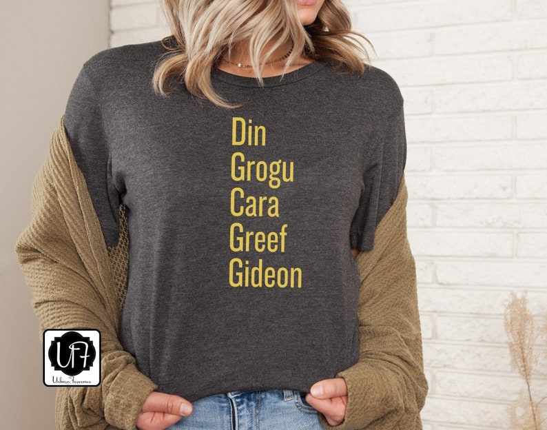 May include: A dark gray t-shirt with the names Din, Grogu, Cara, Greef, and Gideon printed in yellow.