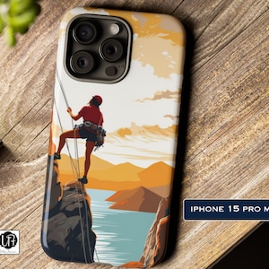 May include: A phone case with a colorful illustration of a person rock climbing on a cliff with a lake and mountains in the background. The text "IPHONE 15 PRO MAX" is at the bottom of the case.