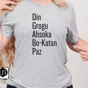 Puede incluir: Camiseta gris con el texto "Din Grogu Ahsoka Bo-Katan Paz" impreso en la parte delantera.
