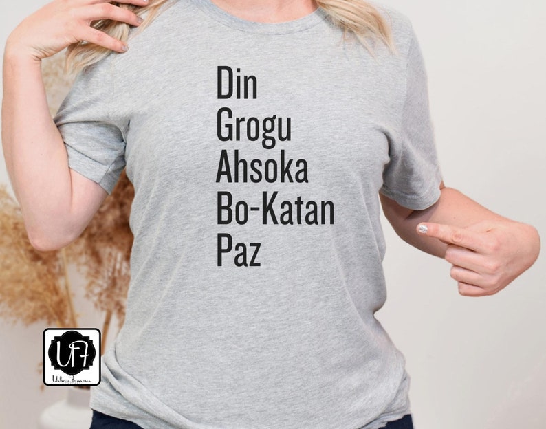 Puede incluir: Camiseta gris con el texto "Din Grogu Ahsoka Bo-Katan Paz" impreso en la parte delantera.