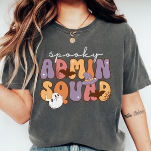 May include: A charcoal gray short-sleeve t-shirt featuring the text "spooky ADMIN SQUAD" in a colorful, wavy font. The graphic includes illustrations of a white ghost, brown bats, a purple witch hat, and spider webs on a dark background.
