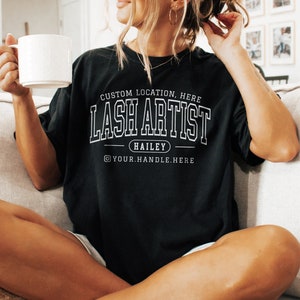 May include: Black t-shirt with white text that reads "CUSTOM LOCATION, HERE LASH ARTIST HAILEY @YOUR.HANDLE.HERE". The model is wearing a size L shirt. The shirt is a casual style.