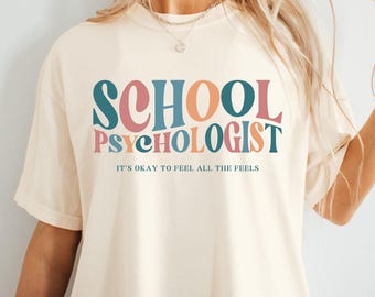 May include: A white t-shirt with the text "SCHOOL PSYCHOLOGIST" in colorful retro lettering. The text "IT'S OKAY TO FEEL ALL THE FEELS" is printed below in a smaller font.