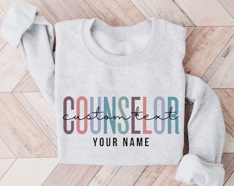 May include: Gray sweatshirt with the text "COUNSELOR" in colorful letters and the words "custom text" and "YOUR NAME" in black script.