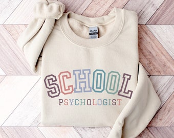 May include: A beige sweatshirt with the text "SCHOOL PSYCHOLOGIST" in rainbow colours. The text is in a retro, collegiate style font.
