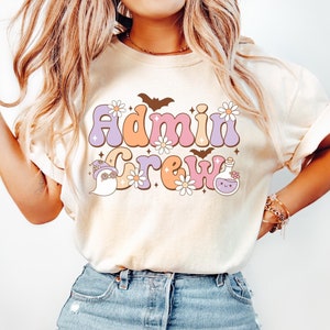 May include: An ivory t-shirt featuring a graphic print with the text "Admin Crew" in a retro font. The design includes illustrations of a ghost wearing a hat, a potion bottle, bats, and daisies in shades of purple, orange, and white.