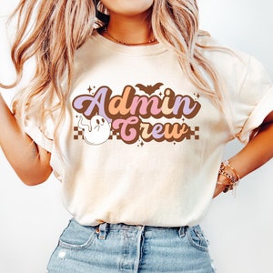 May include: An ivory short-sleeve t-shirt featuring a graphic print with the text Admin Crew in brown, purple, and orange lettering. The design includes a small white ghost, a brown bat, and a checkered pattern border, all centered on the chest.
